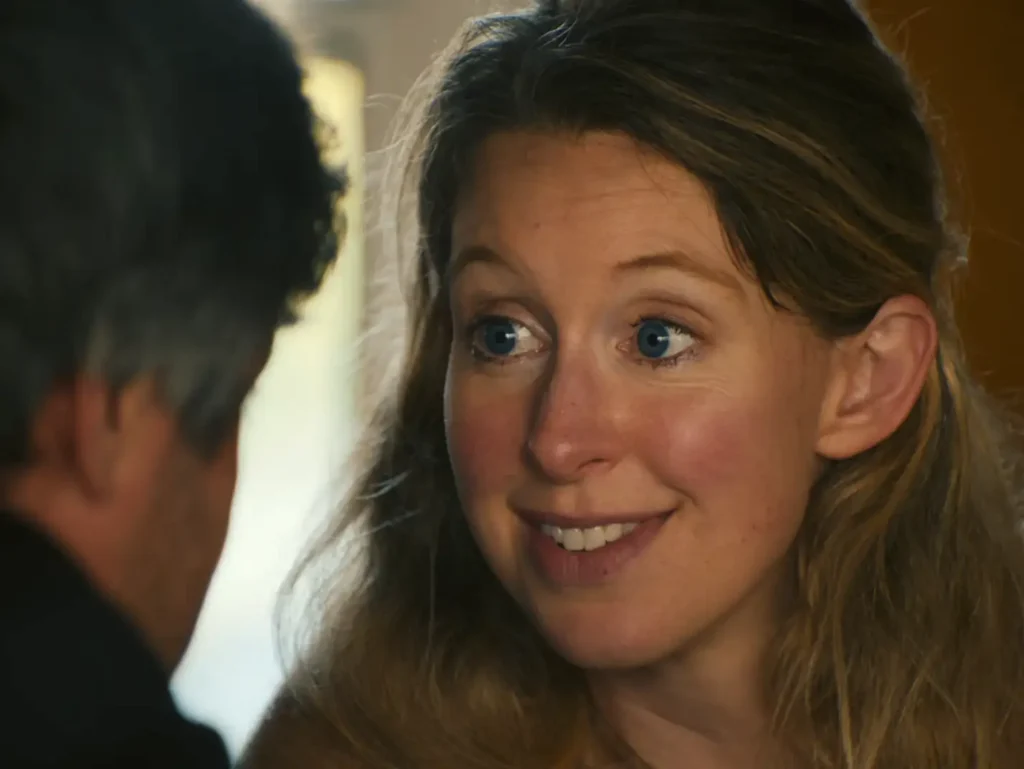 Elizabeth Holmes in a close-up scene from You Can See Everything documentary, smiling during a conversation with Nathan Fielder