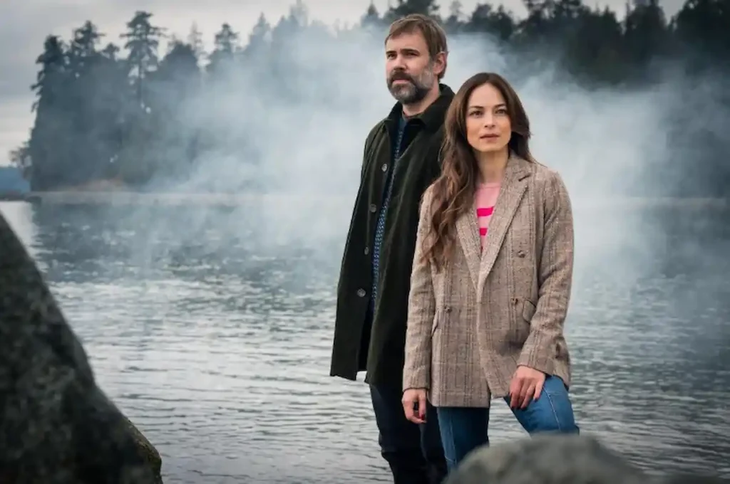 Rossif Sutherland and Kristin Kreuk standing by the waterfront in Murder in a Small Town, set against the misty coastal landscape of Gibsons, British Columbia.