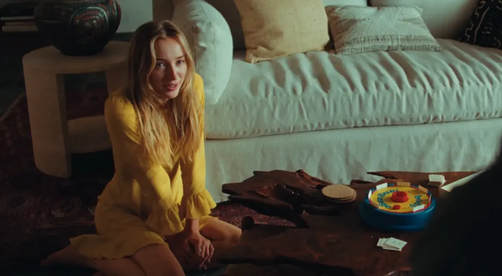 Phoebe Dynevor in a yellow dress sitting on the floor beside a wooden coffee table in a living room, with a white sofa in the background and a colorful board game on the table in Remain movie
